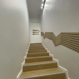 Schody drewniane z szarymi listwami, prowadzące do białych drzwi z trzema poziomymi przeszkleniami, oświetlone listwą LED, z dekoracyjną, drewnianą ścianką po prawej stronie.