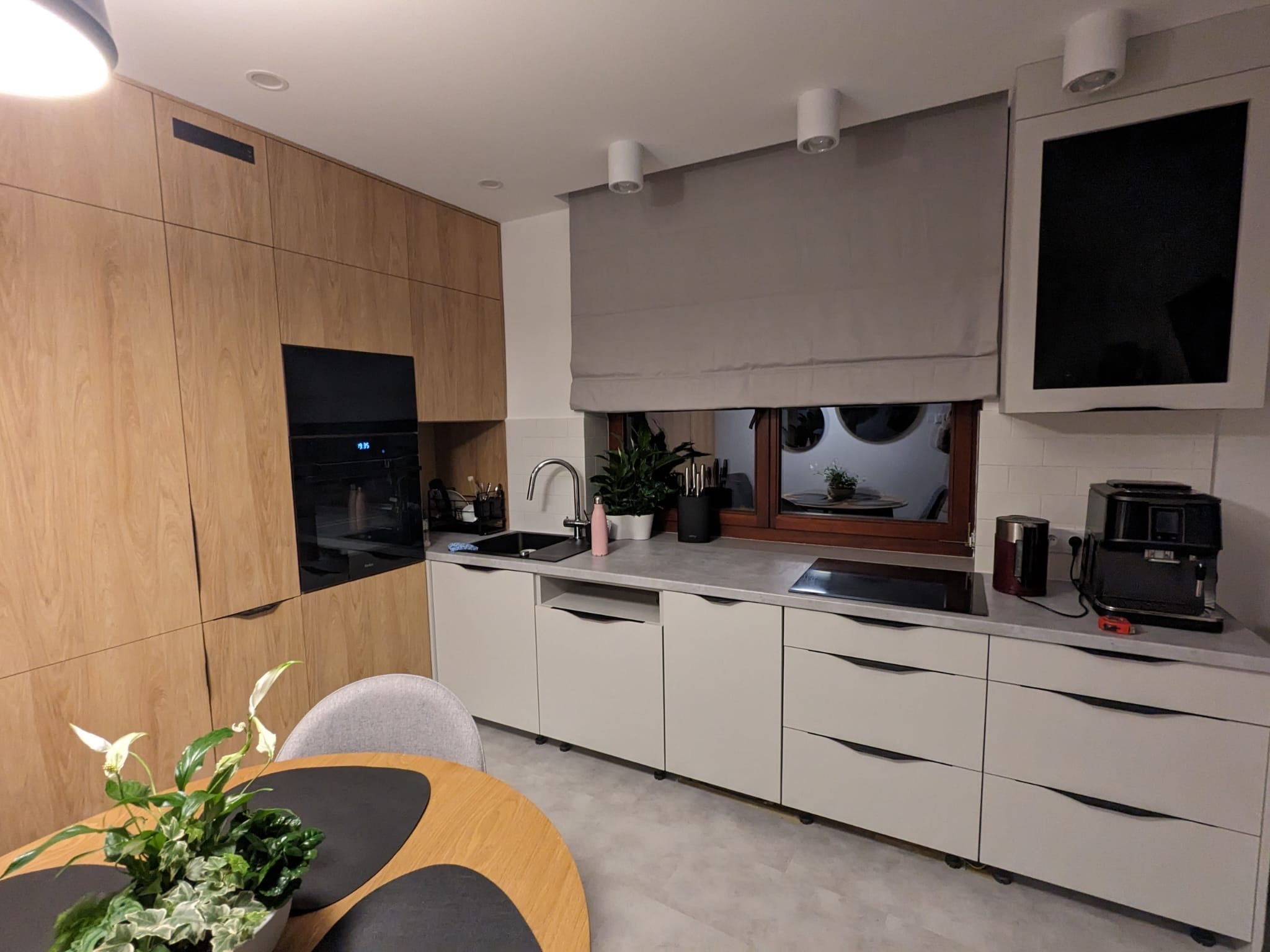 Nowoczesna kuchnia z drewnianymi i białymi szafkami, szarym blatem, płytą indukcyjną, okapem, oraz roślinami w doniczkach na blacie i stole.