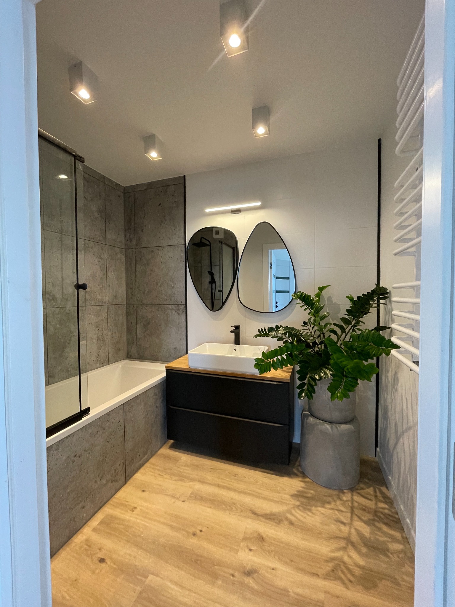 Nowoczesna łazienka z wanną obudowaną szarą płytką, umywalką nablatową z czarną szafką, dwoma asymetrycznymi lustrami i rośliną w doniczce na betonowym postumencie, oświetlona punktowymi lampami...