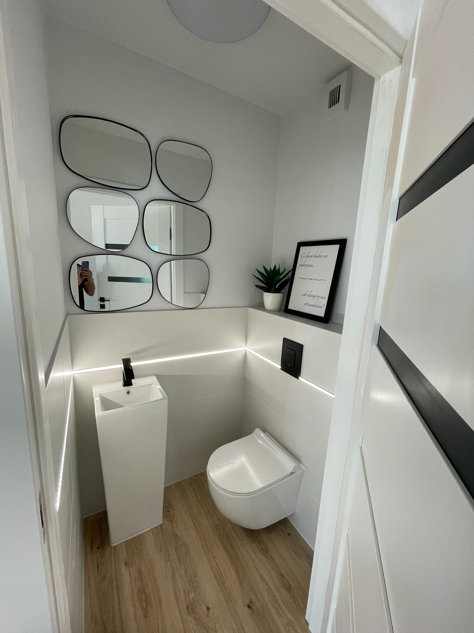 Nowoczesna toaleta w bieli i drewnie: podwieszana muszla, umywalka-monolit z czarną baterią, podświetlenie LED i zestaw luster o organicznych kształtach na ścianie, drewnopodobna podłoga.