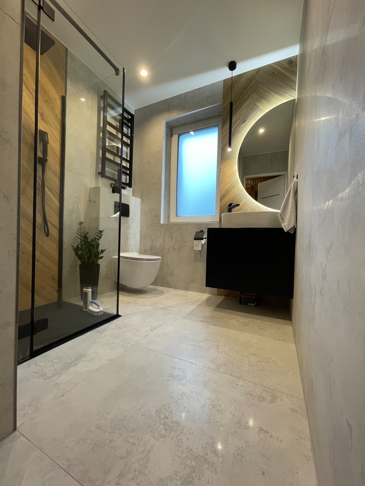 Nowoczesna łazienka z kabiną prysznicową typu walk-in, podwieszaną toaletą, umywalką z szafką i okrągłym lustrem z podświetleniem LED, wykończona w szarościach z drewnianymi akcentami.