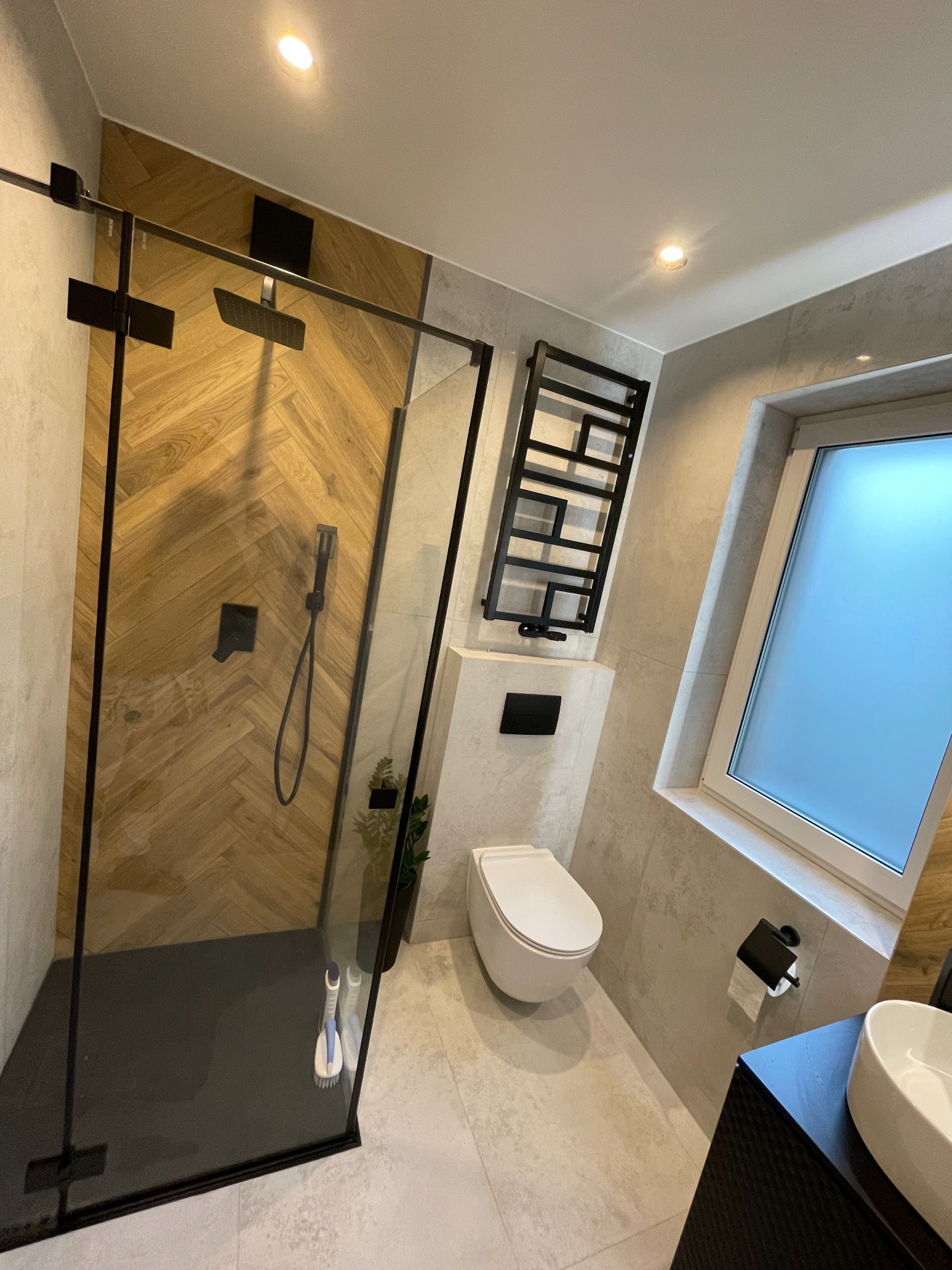 Nowoczesna łazienka z kabiną prysznicową typu walk-in, wykończoną drewnem ułożonym w jodełkę, czarną armaturą i grzejnikiem drabinkowym, jasnymi płytkami i toaletą podwieszaną.