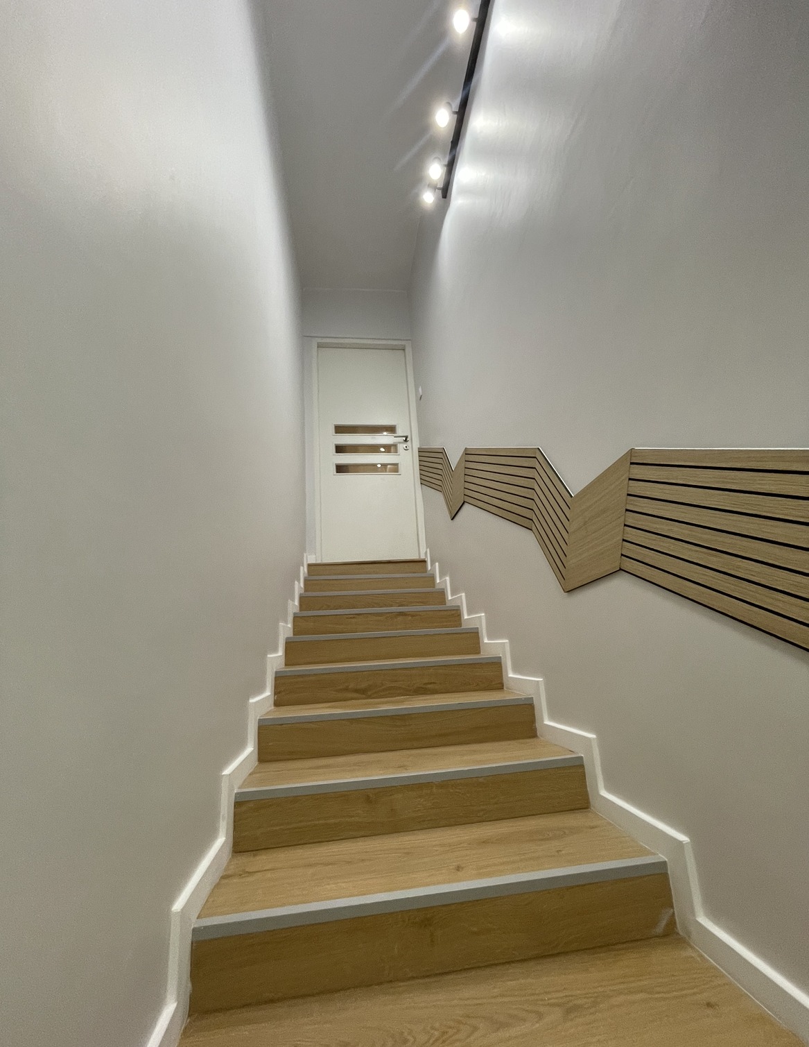 Schody drewniane z szarymi listwami, prowadzące do białych drzwi z trzema poziomymi przeszkleniami, oświetlone listwą LED, z dekoracyjną, drewnianą ścianką po prawej stronie.