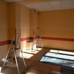 DIERO - Pokój w trakcie odświeżania: ściany pomalowane na pomarańczowo z bordowym pasem, dwie drabiny aluminiowe, wiadro z farbą i zabezpieczona podłoga folią malarską i kartonami.