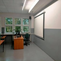 DIERO - Klasa po odświeżeniu: ściany pomalowane na biało i szaro, widoczny ekran projekcyjny zwinięty do góry, biurko nauczycielskie z komputerem i rośliną, krzesła ustawione na stołach.