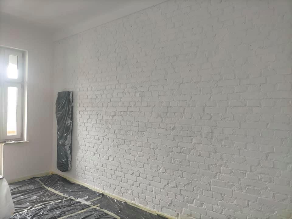 Biała, pomalowana ściana z cegły w pokoju, zabezpieczona folią podłoga, okno z lewej strony, grzejnik owinięty folią.