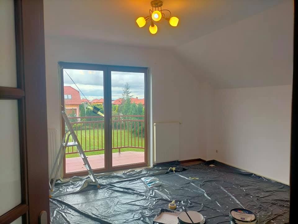 Pokój przygotowany do malowania: zabezpieczona folią podłoga, drabina oparta o okno z widokiem na ogród, wiaderka z farbą Dulux i kuweta malarska.