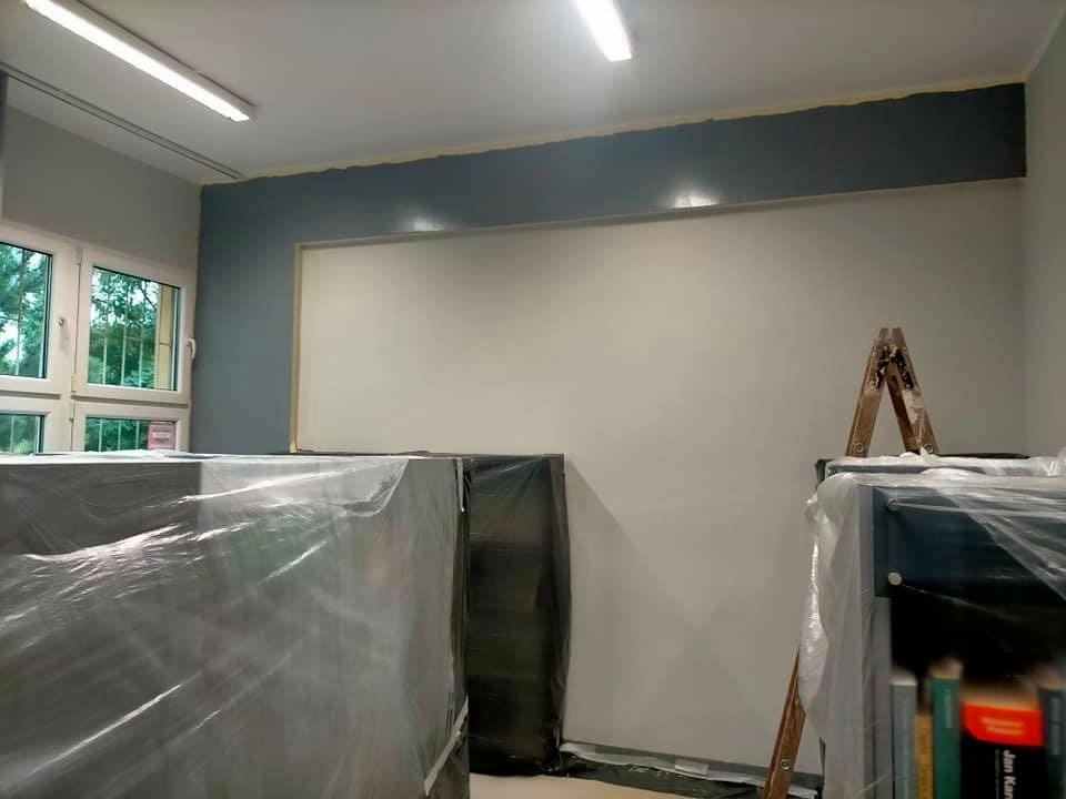 Pokój w trakcie odświeżania: fragment ściany pomalowany na szaro-niebiesko z taśmą malarską, reszta ściany biała, zabezpieczone folią meble i drabina oparta o regał.