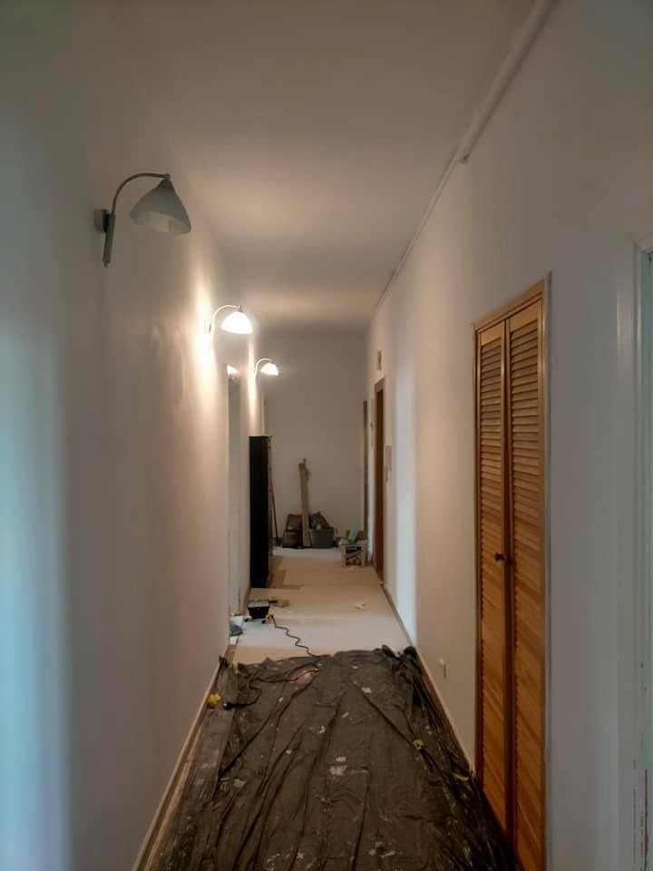 Korytarz w trakcie odświeżania ścian, widoczne lampy ścienne, zabezpieczona podłoga folią malarską, ślady po pracach na ścianach.