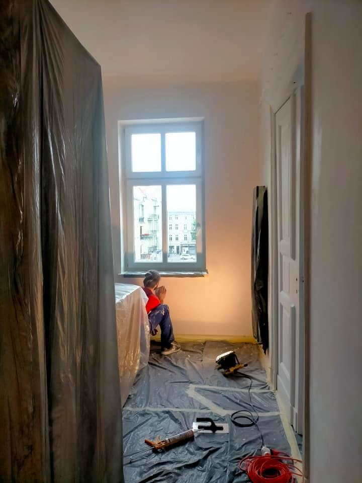 Pokój w trakcie odświeżania: malarz siedzi przy oknie w zabezpieczonym pomieszczeniu, widoczne narzędzia i lampa na folii ochronnej.