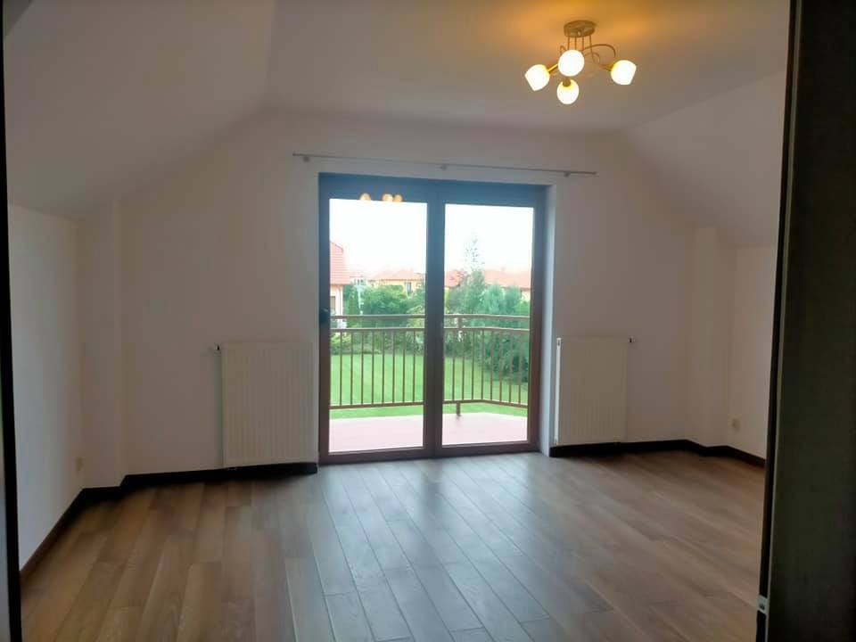 Puste poddasze z drewnianą podłogą i balkonem z widokiem na ogród, białe ściany i grzejniki, sufit z lampą w stylu retro.