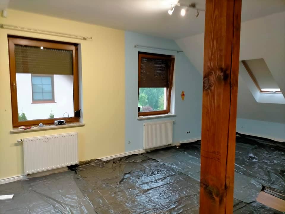 Pokój w trakcie przygotowań do malowania: zabezpieczona folią podłoga, pomalowane fragmenty ścian na żółto i niebiesko, widoczny drewniany słup konstrukcyjny.
