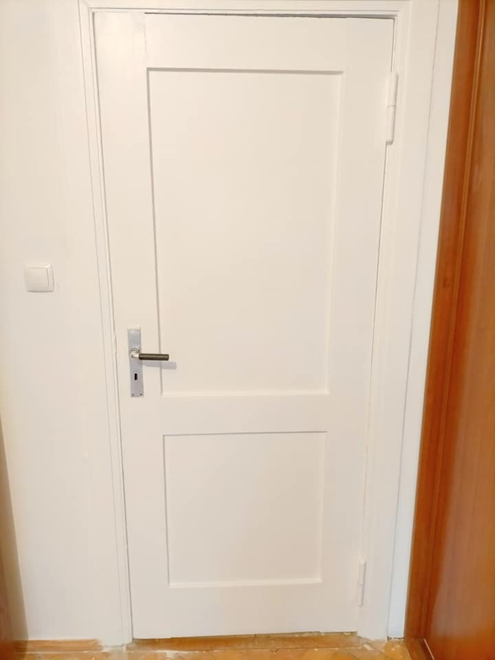 Białe, pomalowane drzwi wewnętrzne z prostokątnymi panelami, srebrna klamka, widoczne ślady po malowaniu przy podłodze.