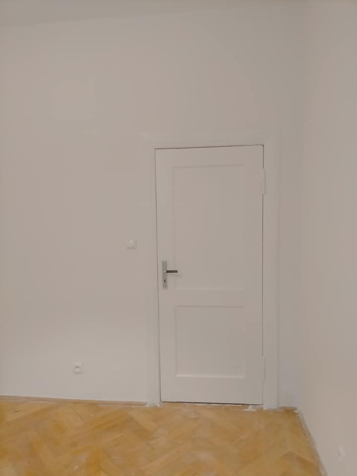 Wnętrze pokoju po remoncie: ściany pomalowane na biało, białe drzwi z srebrną klamką oraz parkiet ułożony w jodełkę.