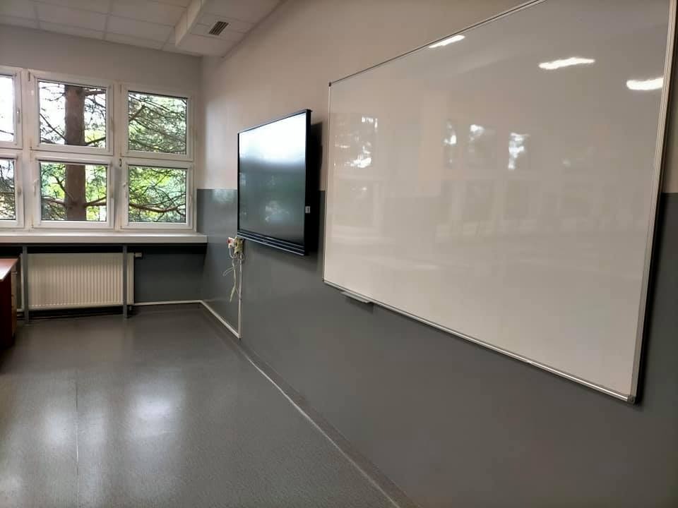Wnętrze pomalowanej na szaro sali lekcyjnej z białymi ścianami u góry, z oknem z widokiem na drzewa, tablicą interaktywną i białą tablicą.