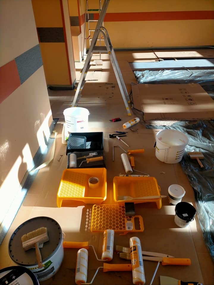 Przygotowane narzędzia malarskie: wałki, pędzle, kuwety, drabina i farby w wiadrach na podłodze zabezpieczonej kartonem i folią, w pomieszczeniu z częściowo pomalowanymi ścianami.