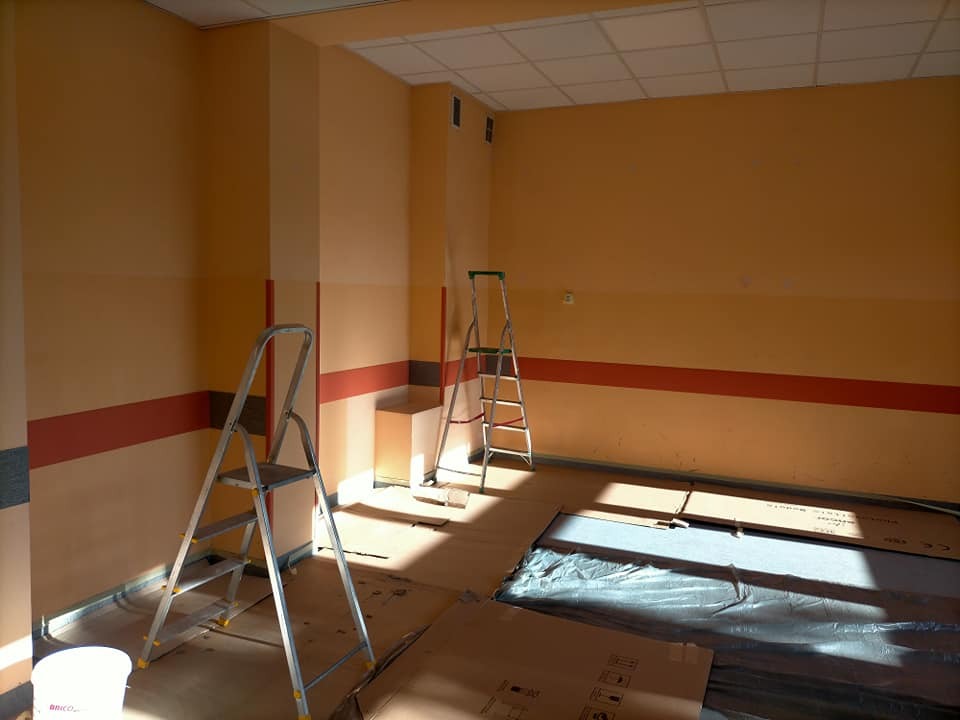 Pokój w trakcie odświeżania: ściany pomalowane na pomarańczowo z bordowym pasem, dwie drabiny aluminiowe, wiadro z farbą i zabezpieczona podłoga folią malarską i kartonami.