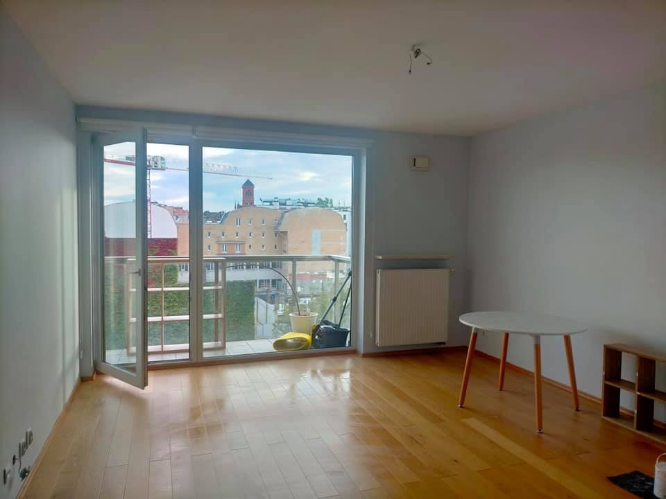 Pusty pokój z parkietem, białymi ścianami i otwartym balkonem z widokiem na miasto, widoczne ślady po malowaniu na ścianach, brak mebli poza stołem i regałem.