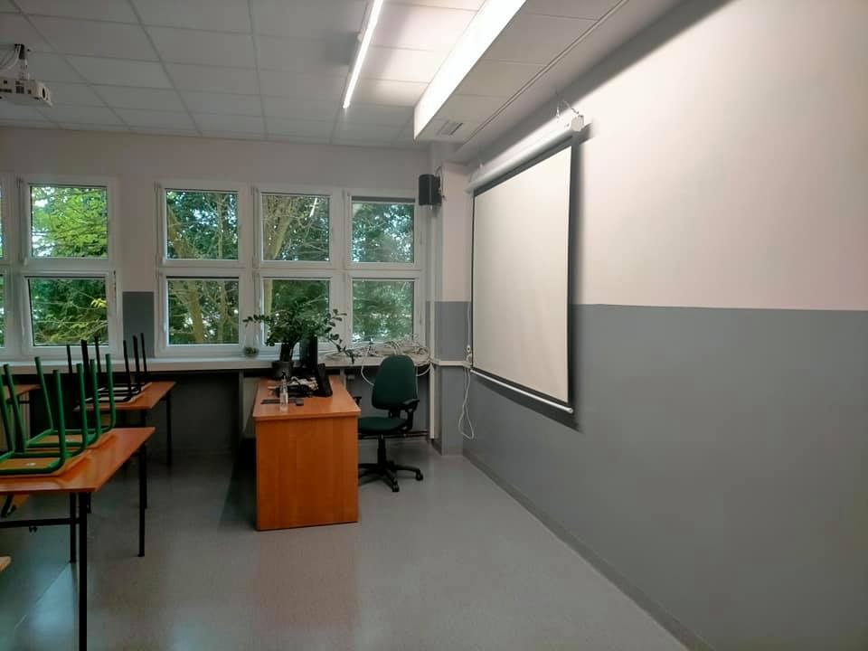 Klasa po odświeżeniu: ściany pomalowane na biało i szaro, widoczny ekran projekcyjny zwinięty do góry, biurko nauczycielskie z komputerem i rośliną, krzesła ustawione na stołach.