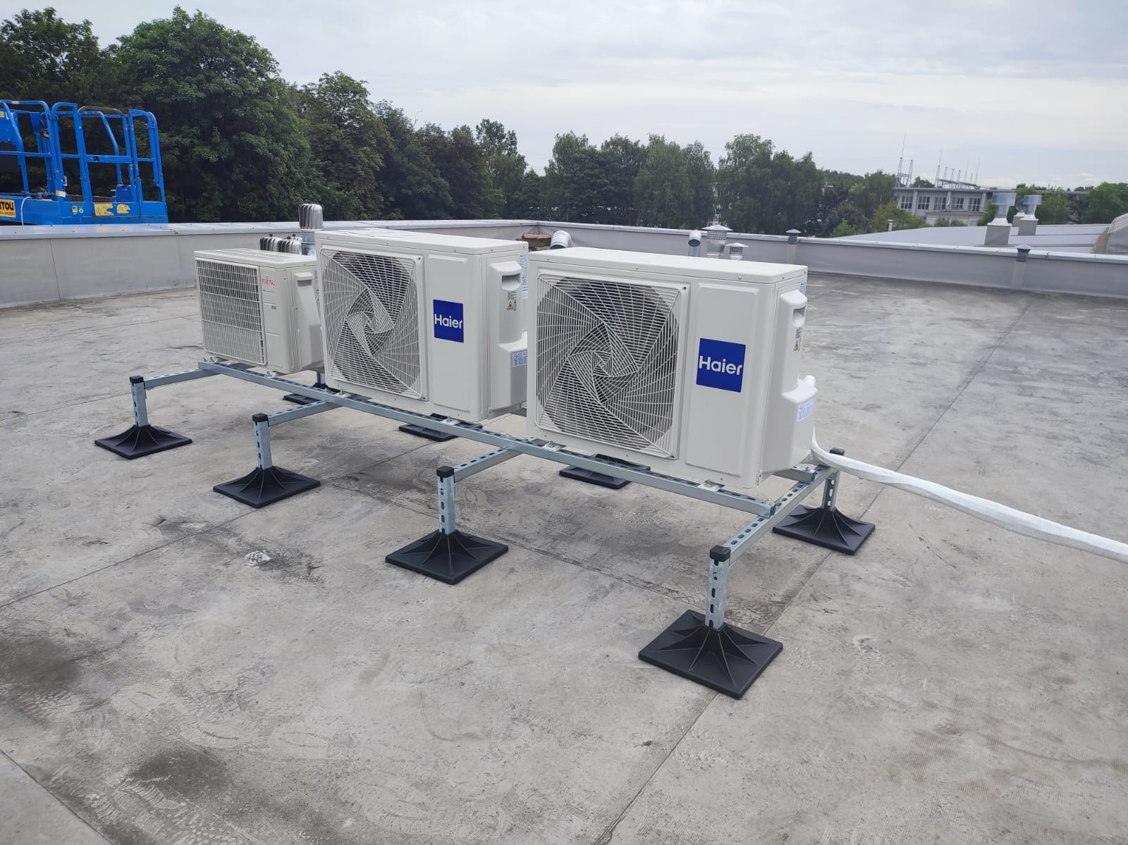 Instalacja kilku jednostek zewnętrznych klimatyzacji Haier i Fujitsu na płaskim dachu, widoczny niebieski podnośnik koszowy w oddali.