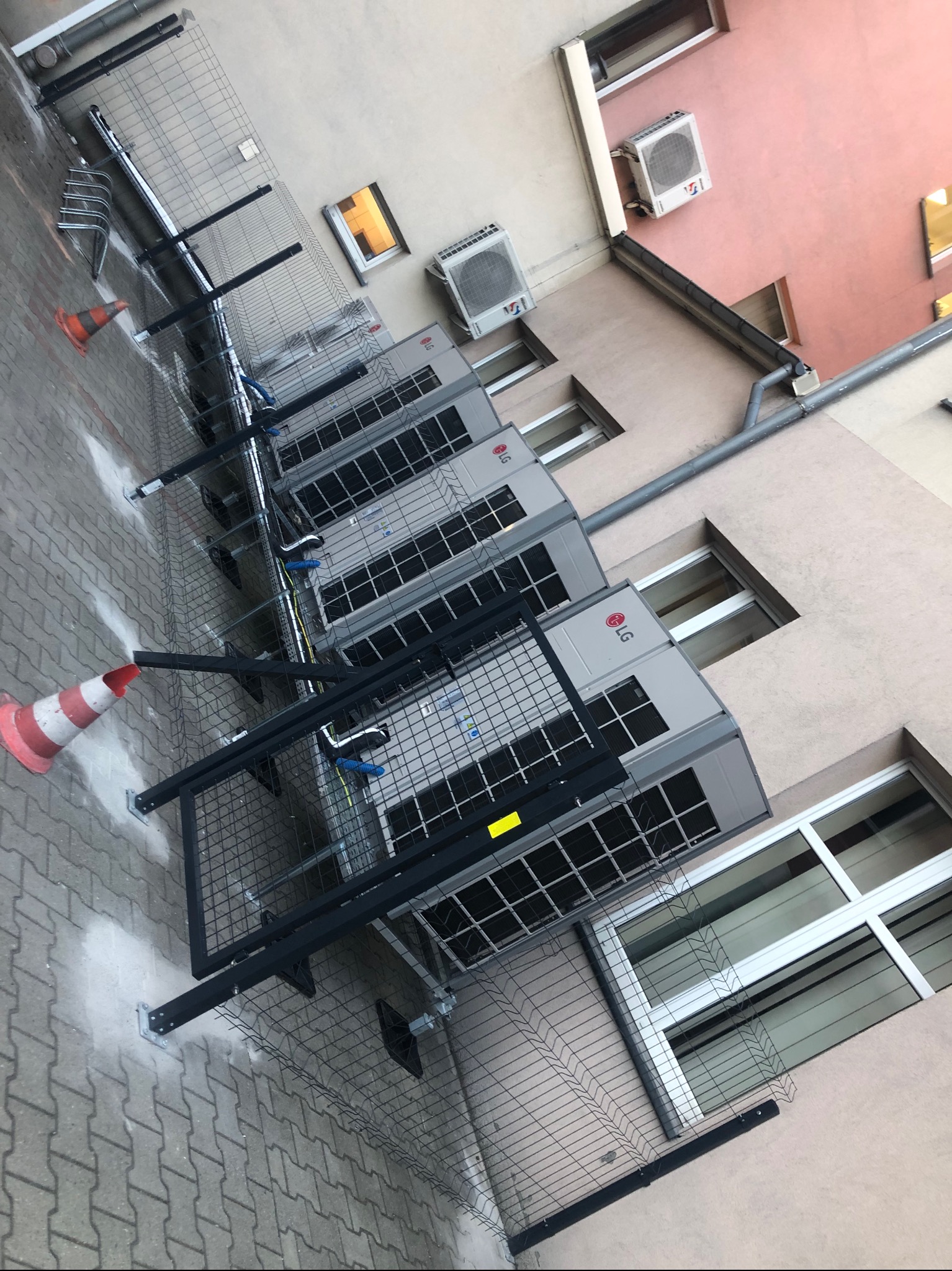 Rząd zewnętrznych jednostek klimatyzacji LG zamontowanych na ścianie budynku, zabezpieczonych czarną metalową konstrukcją, z pomarańczowo-białym pachołkiem na chodniku z kostki brukowej.