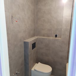 Paweł Spiączka - Szara łazienka w trakcie remontu: wisząca toaleta, płytki imitujące beton, wystające przewody elektryczne i puszka pod gniazdko.