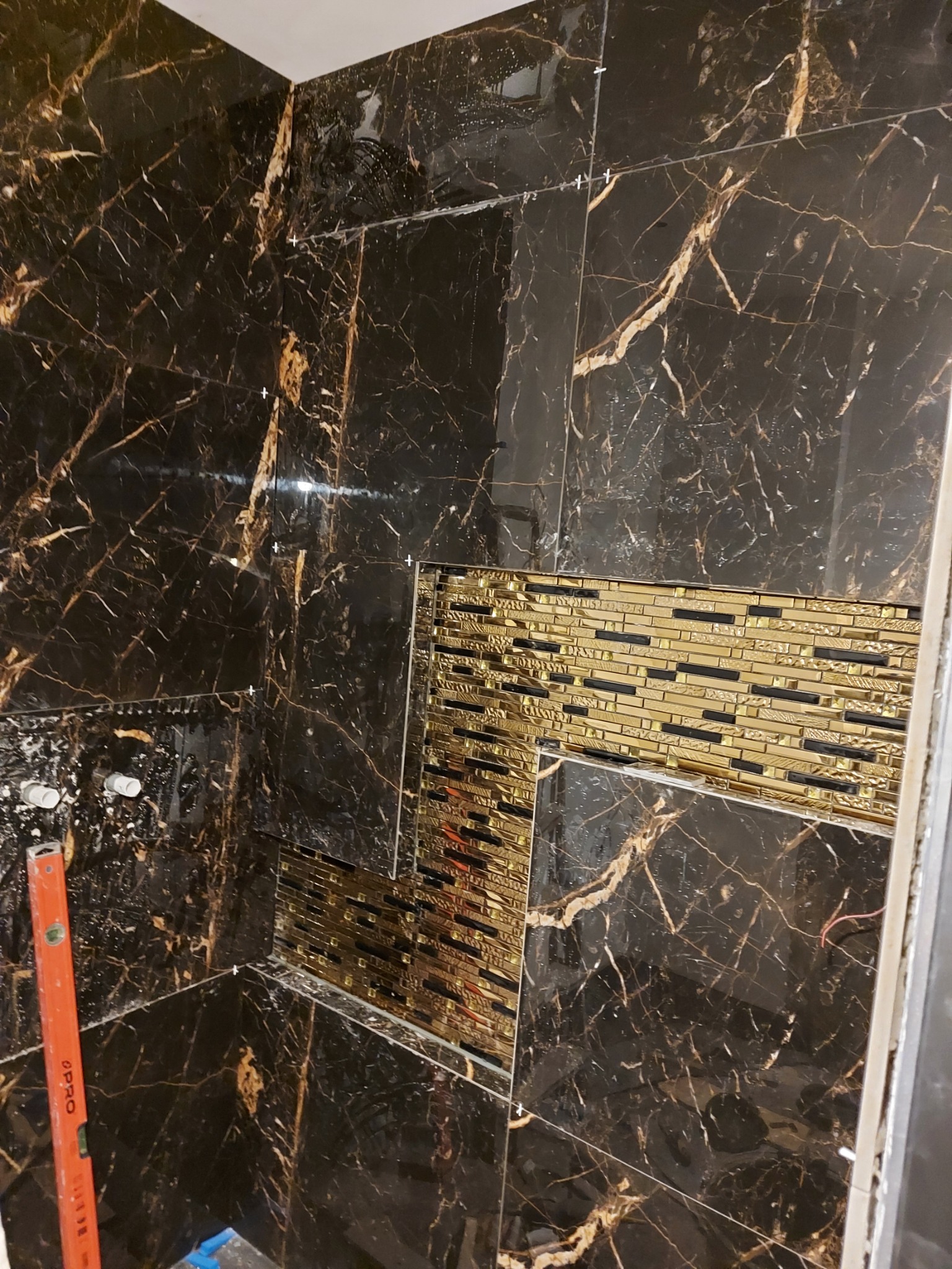 Montaż czarnych płytek imitujących marmur z złotymi żyłami oraz dekoru z mozaiki w łazience, widoczna poziomica i białe rurki.