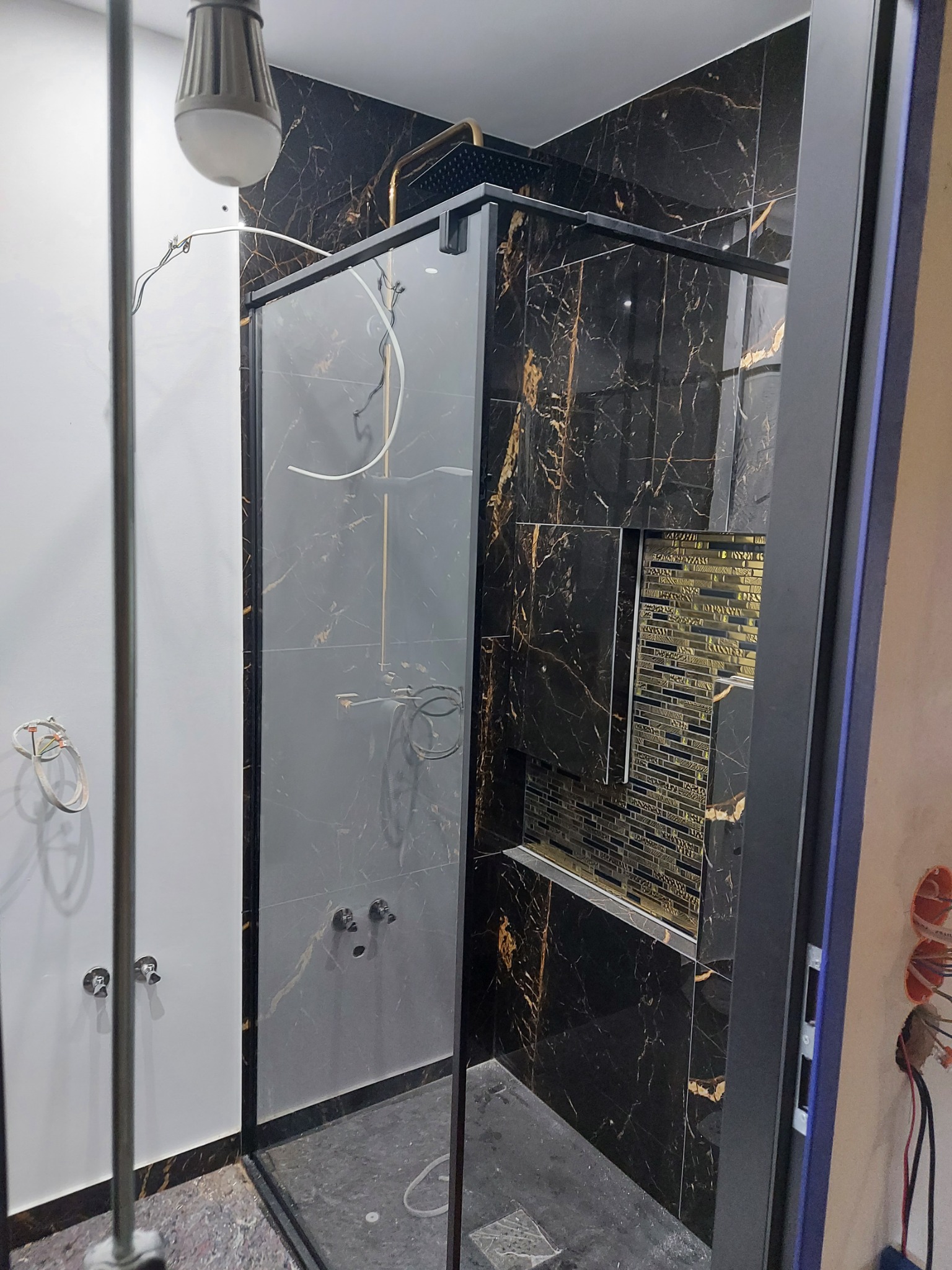 Nowoczesna kabina prysznicowa z czarną marmurową okładziną i złotymi akcentami, widoczne niedokończone prace elektryczne na ścianie.