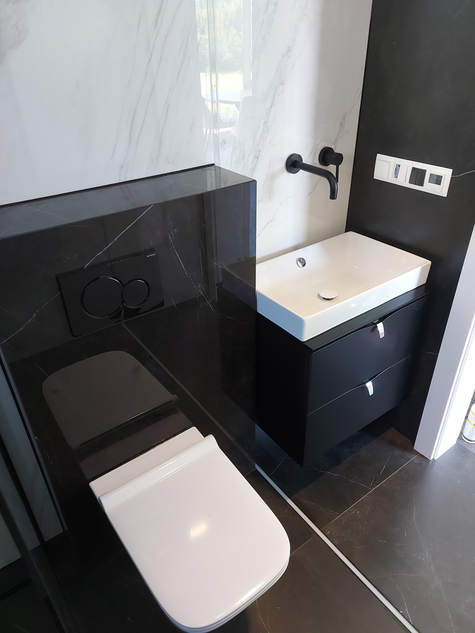 Nowoczesna łazienka w czarno-białej kolorystyce: wisząca toaleta z czarną spłuczką Geberit, umywalka nablatowa na czarnej szafce z chromowanymi uchwytami, czarna bateria ścienna i marmurowe płytki...