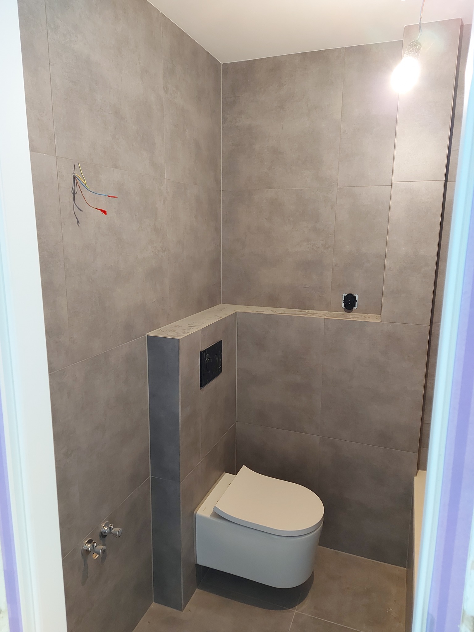 Szara łazienka w trakcie remontu: wisząca toaleta, płytki imitujące beton, wystające przewody elektryczne i puszka pod gniazdko.