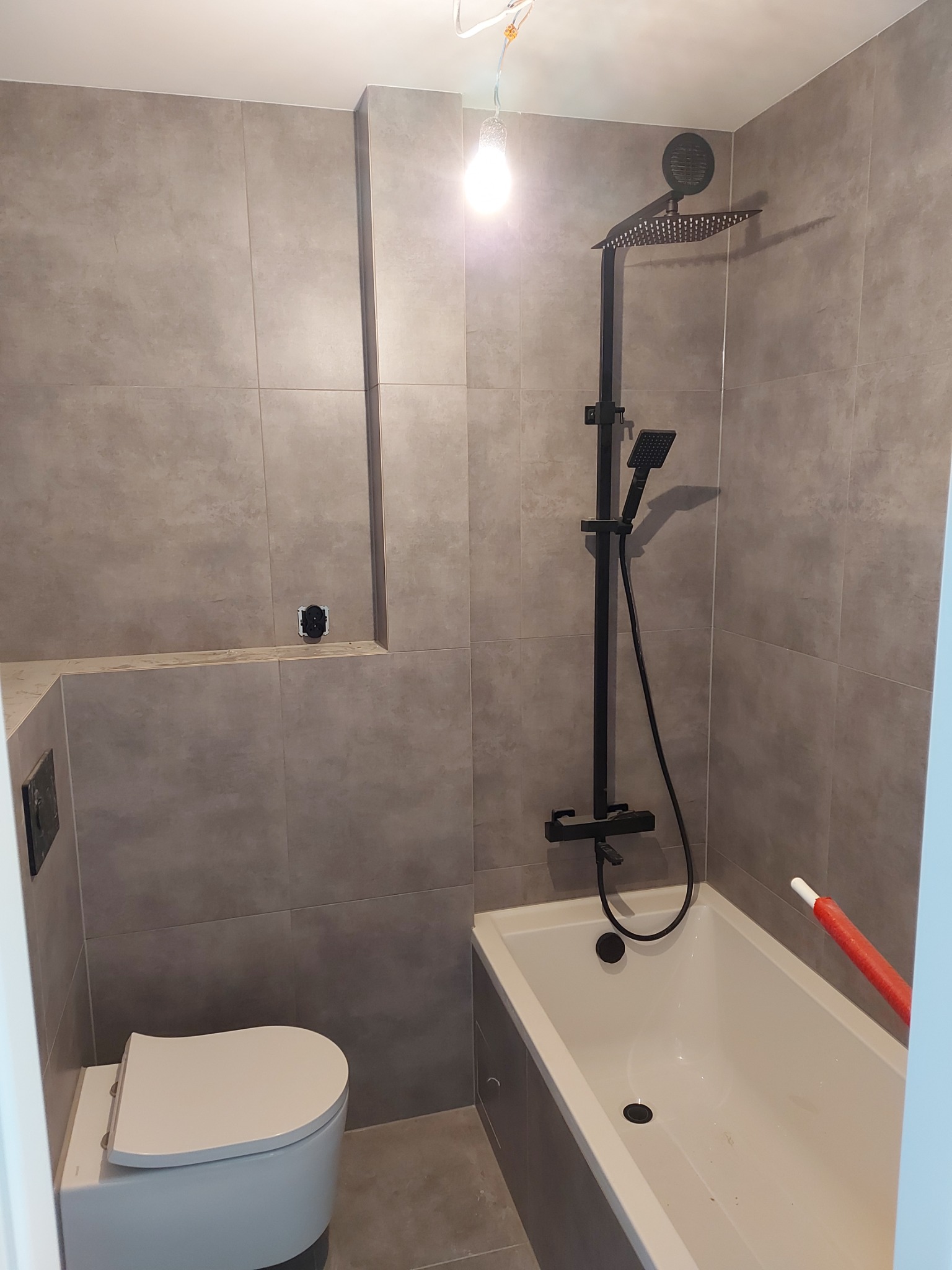 Wykończona łazienka z szarymi płytkami, białą wanną i toaletą oraz czarną armaturą prysznicową; widoczne niedokończone podłączenie elektryczne na suficie.