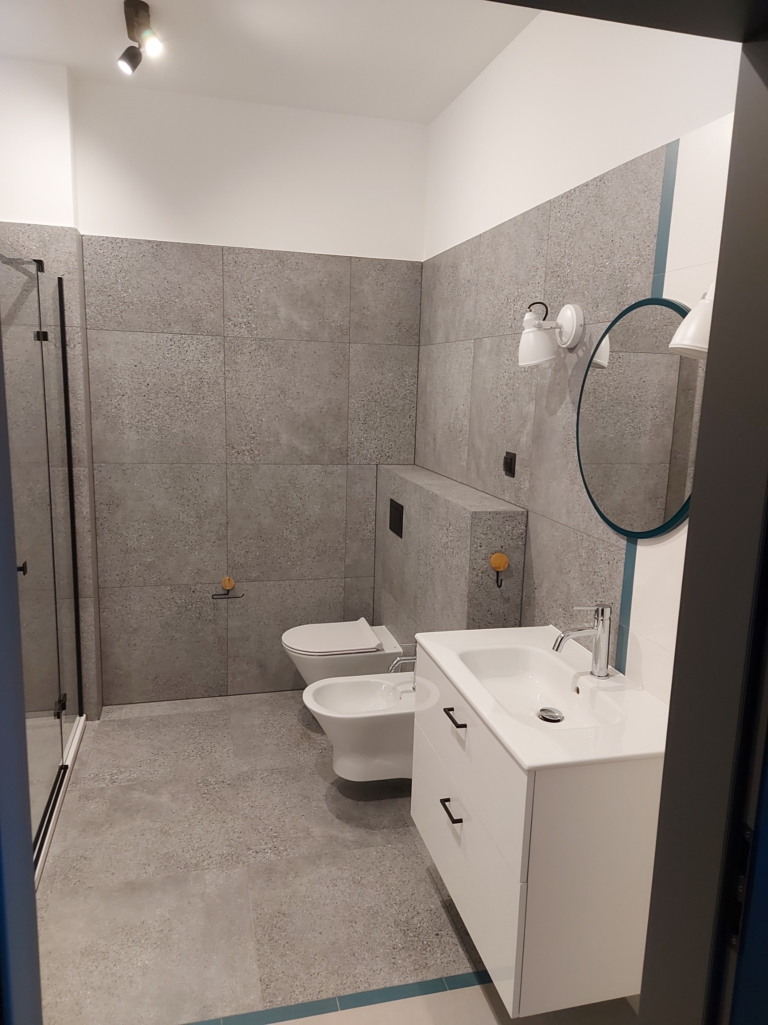 Nowoczesna łazienka z szarymi płytkami imitującymi lastryko, białą armaturą i szafką pod umywalką z czarnymi uchwytami.