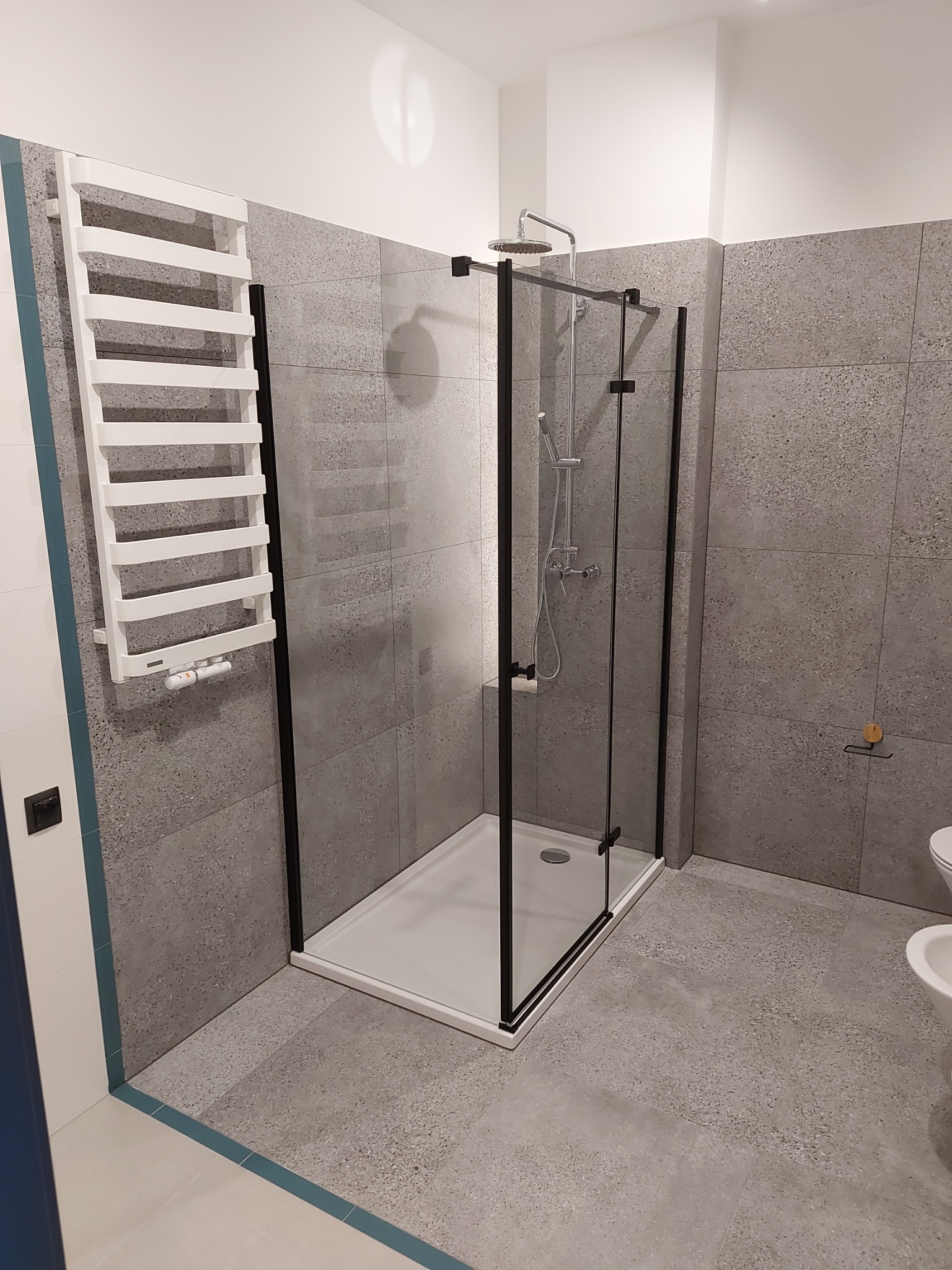 Nowoczesna kabina prysznicowa z czarnymi profilami i szarymi płytkami w łazience po remoncie w Bytomiu. Na ścianie biały grzejnik drabinkowy.