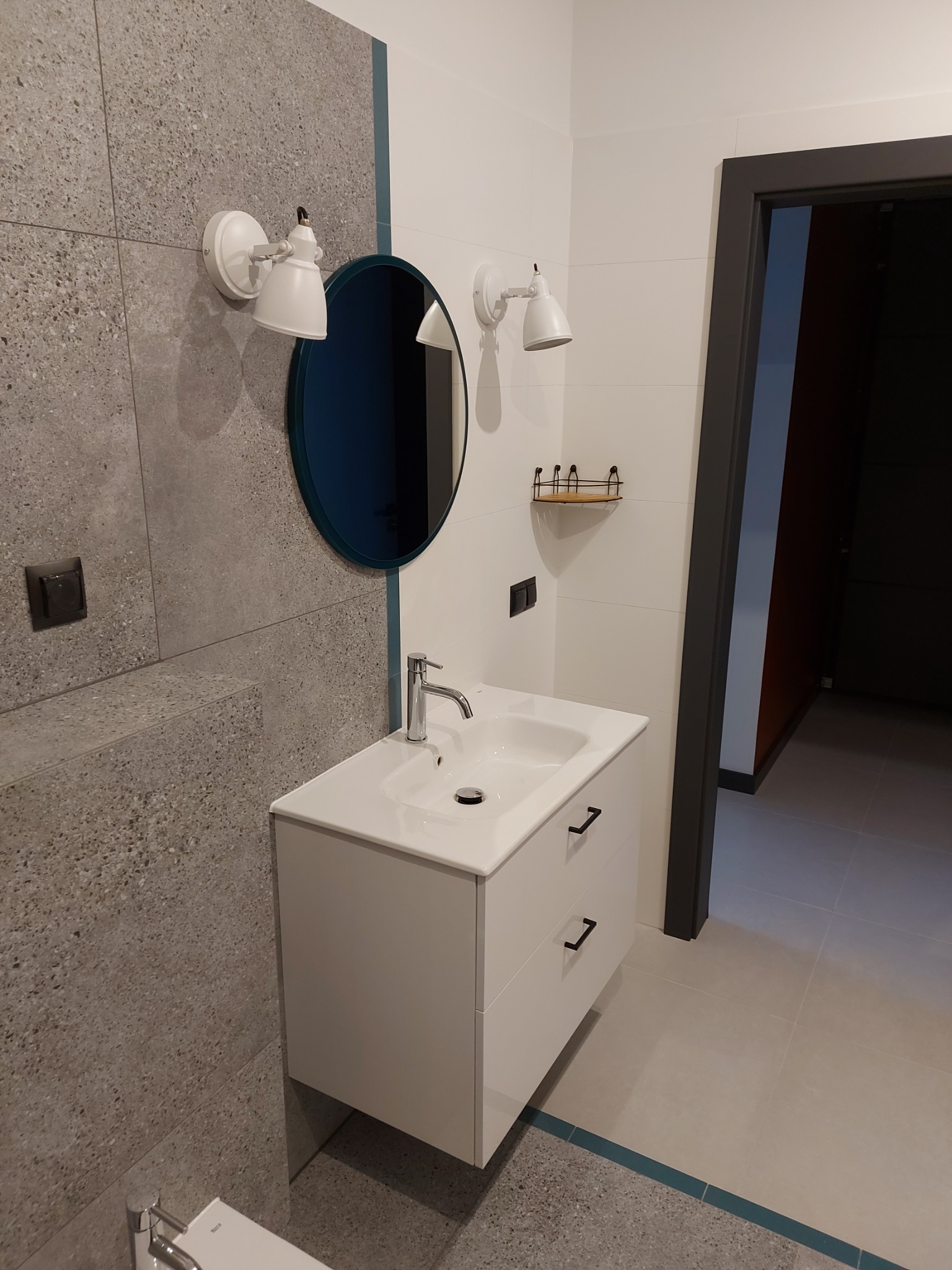Nowoczesna łazienka z umywalką nablatową na białej szafce, okrągłym lustrem w niebieskiej ramie i szarymi płytkami imitującymi kamień na ścianie, oświetlona dwoma białymi kinkietami.