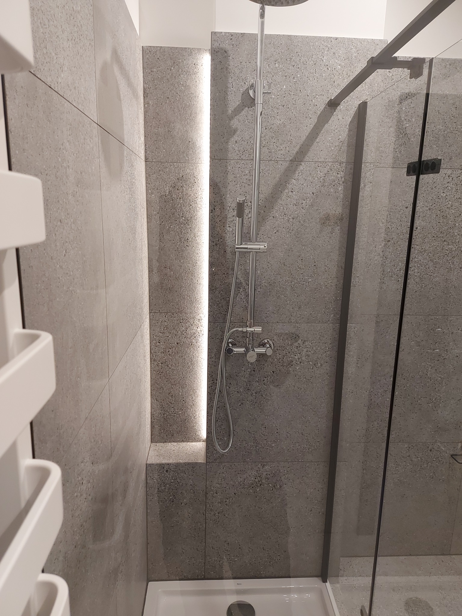 Nowoczesna łazienka z szarymi płytkami imitującymi kamień, kabina prysznicowa z czarnymi okuciami, podświetlana wnęka i biały grzejnik drabinkowy.