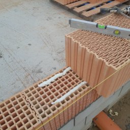 DOMY ENERGOOSZCZĘDNE-JUWO POROTON SPOLKA Z O.O - Wznoszenie ściany z pustaków ceramicznych z użyciem pianki montażowej, poziomicy i sznurka murarskiego, widoczna paleta z pustakami w tle.