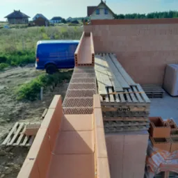 Wznoszenie ścian domu z czerwonej ceramiki poryzowanej, widoczne palety z pustakami Juwo oraz niebieski bus na tle krajobrazu podmiejskiego.