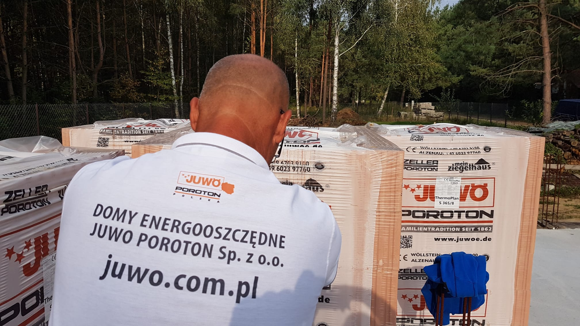 Pracownik w koszulce z logo producenta materiałów budowlanych Juwo Poroton ogląda palety z cegłami Porotherm na placu budowy, w tle las.