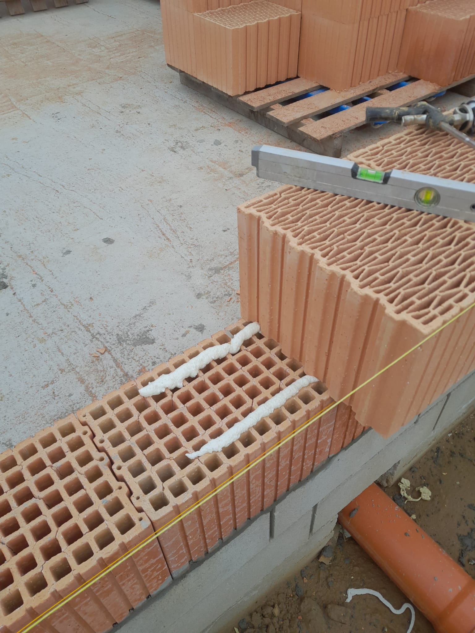 Wznoszenie ściany z pustaków ceramicznych z użyciem pianki montażowej, poziomicy i sznurka murarskiego, widoczna paleta z pustakami w tle.