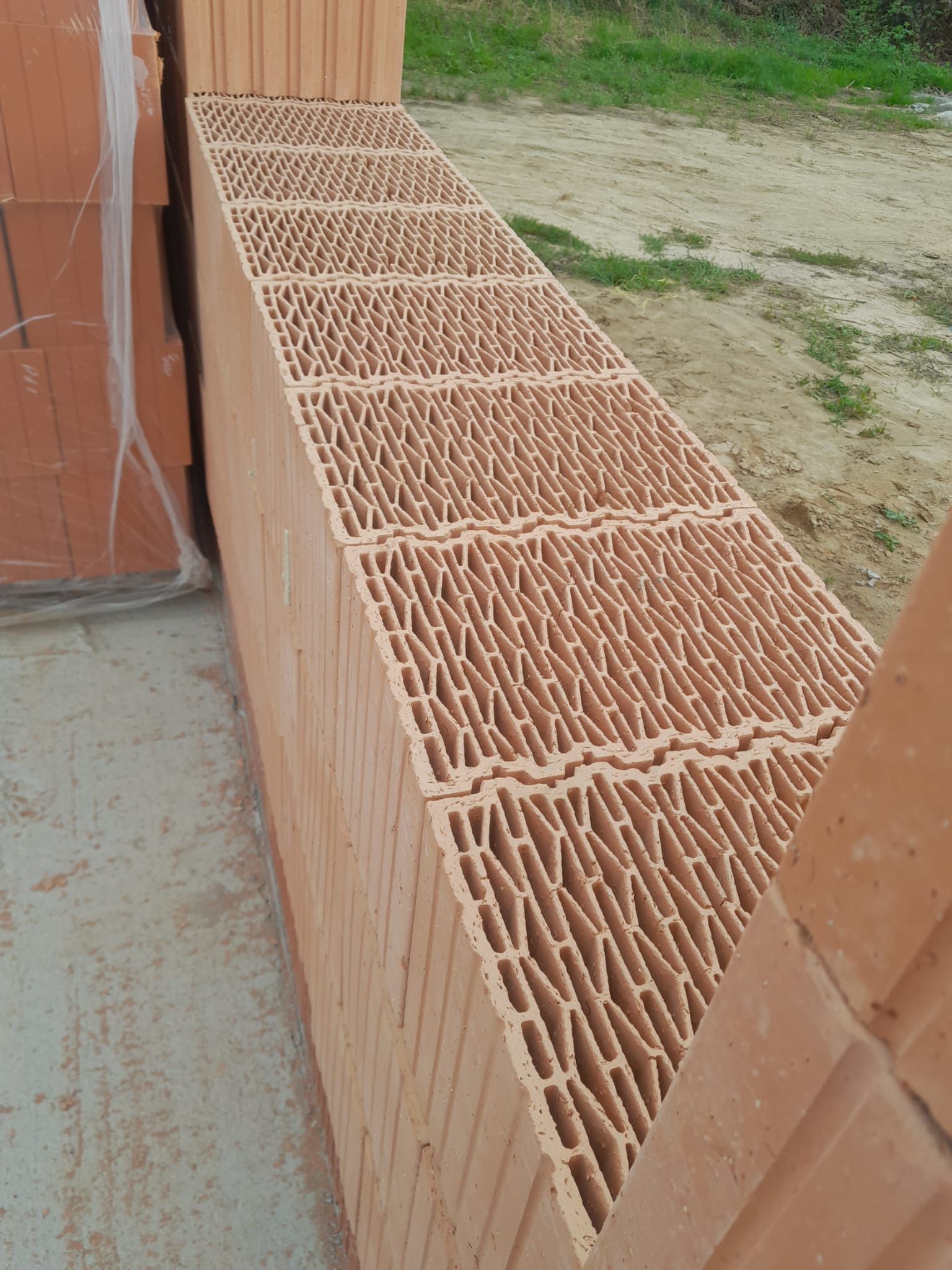 Ułożone bloczki ceramiczne z widoczną strukturą wewnętrzną, część zafoliowana, na betonowej powierzchni, w tle ziemia z zielenią.