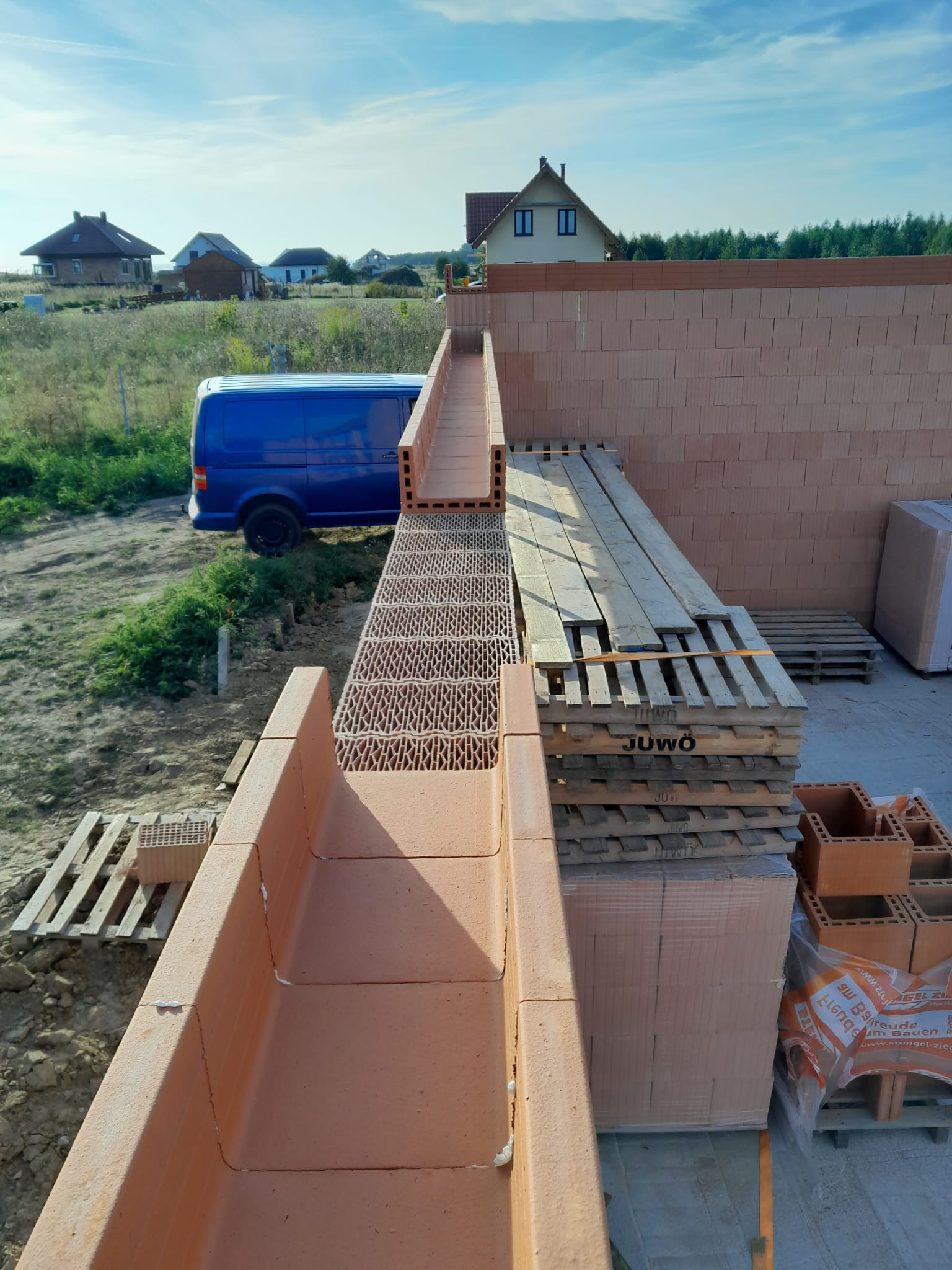 Wznoszenie ścian domu z czerwonej ceramiki poryzowanej, widoczne palety z pustakami Juwo oraz niebieski bus na tle krajobrazu podmiejskiego.