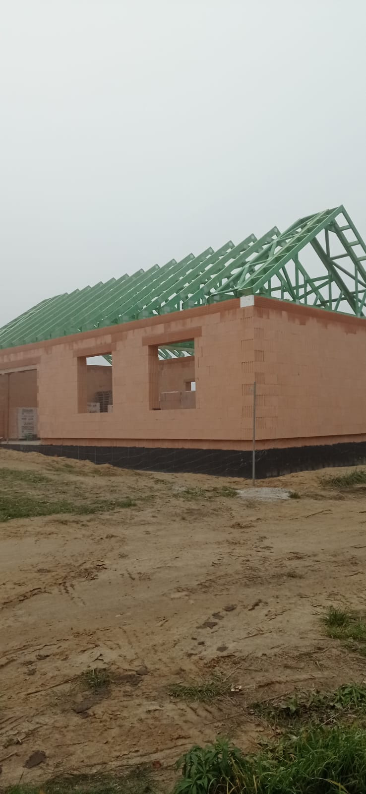 Surowy stan domu jednorodzinnego z cegły ceramicznej z zieloną więźbą dachową, widok z zewnątrz na tle pochmurnego nieba.