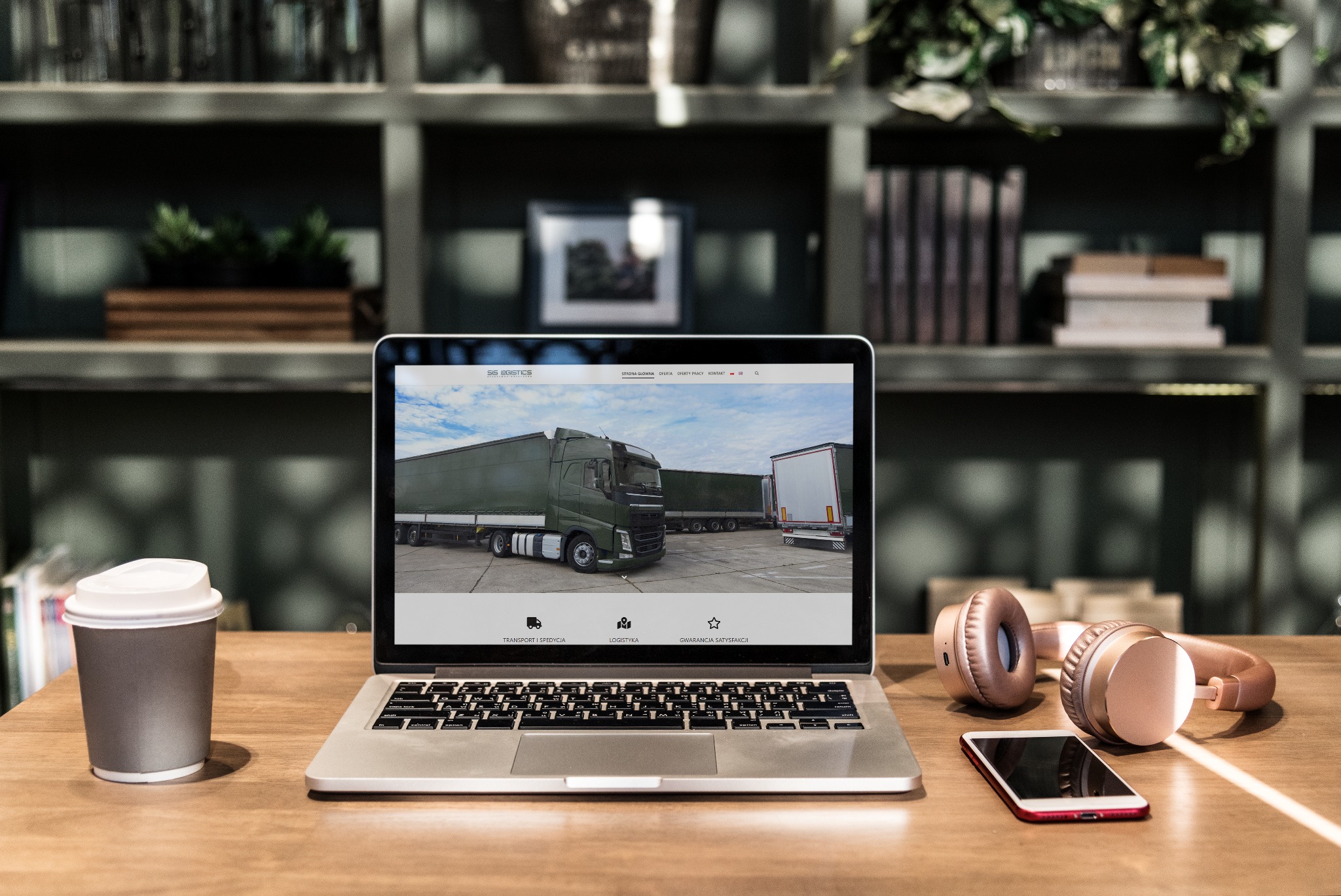Laptop wyświetlający stronę internetową firmy transportowej, obok słuchawki, smartfon i kawa na biurku na tle regału z książkami.