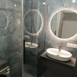 Nowoczesna łazienka z kabiną prysznicową, umywalką nablatową na ciemnej szafce i okrągłym lustrem z podświetleniem LED na ścianie wykończonej płytkami imitującymi marmur.