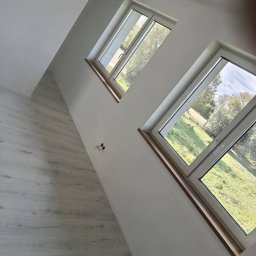 MANIA-BUD usługi remontowo budowlane - Jasne panele podłogowe i dwa okna z drewnianymi parapetami w nowym wnętrzu w Warszawie. Świeżo pomalowane ściany i widok na zieleń za oknem.
