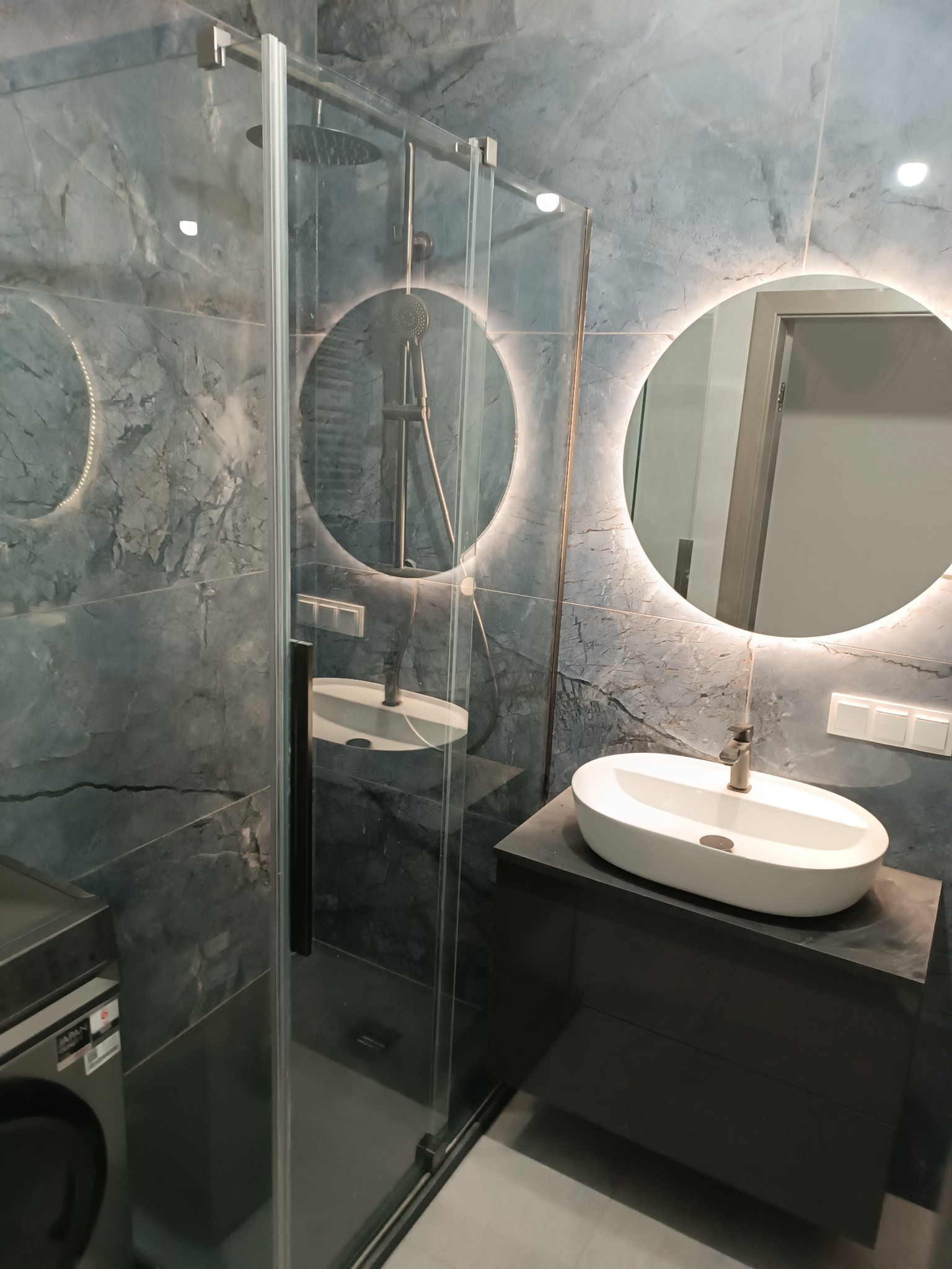 Nowoczesna łazienka z kabiną prysznicową, umywalką nablatową na ciemnej szafce i okrągłym lustrem z podświetleniem LED na ścianie wykończonej płytkami imitującymi marmur.