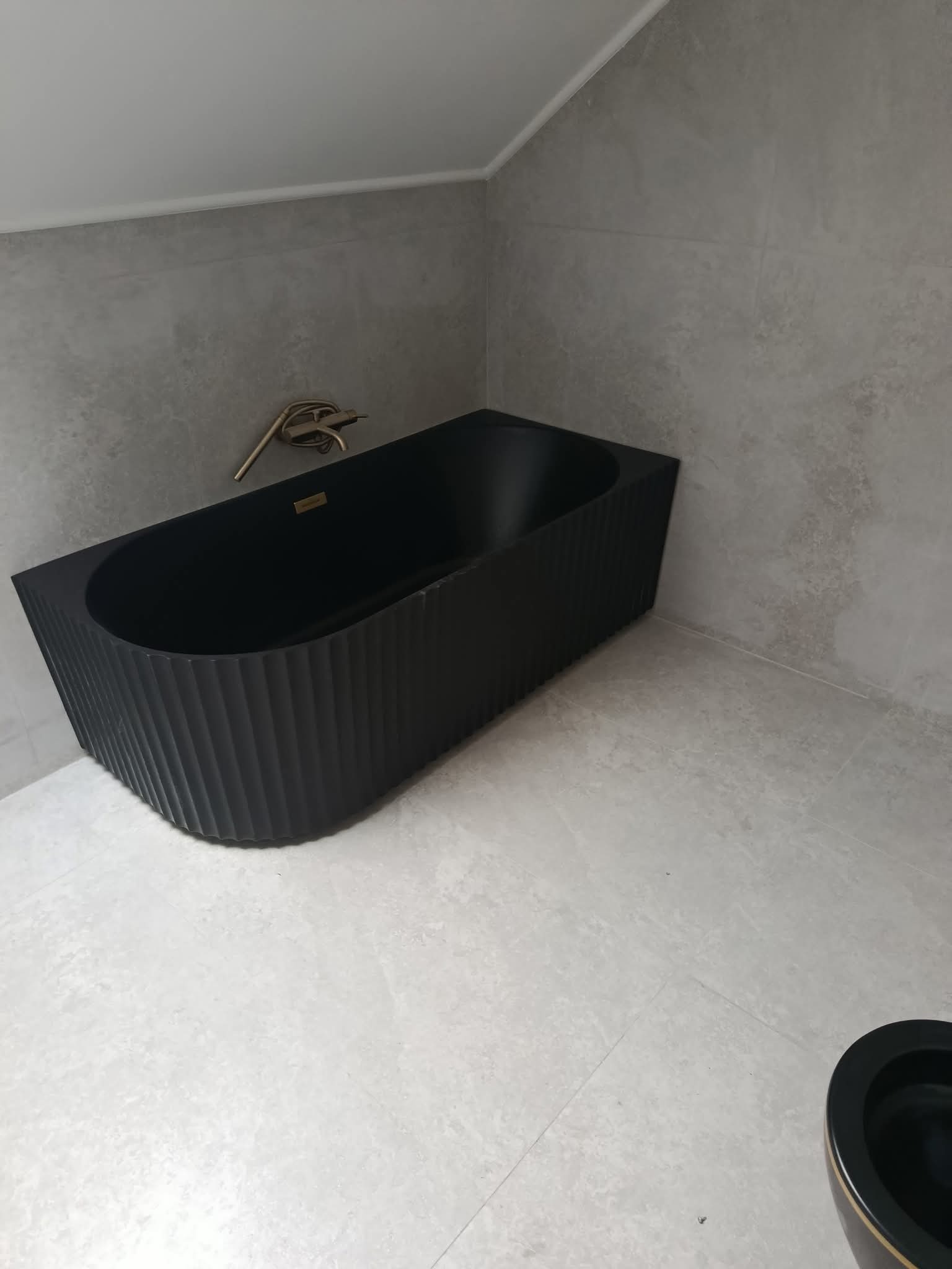 Nowoczesna, czarna wanna z żłobieniami i złotą armaturą w łazience wykończonej szarymi płytkami. Minimalistyczny design, ukończony remont łazienki.