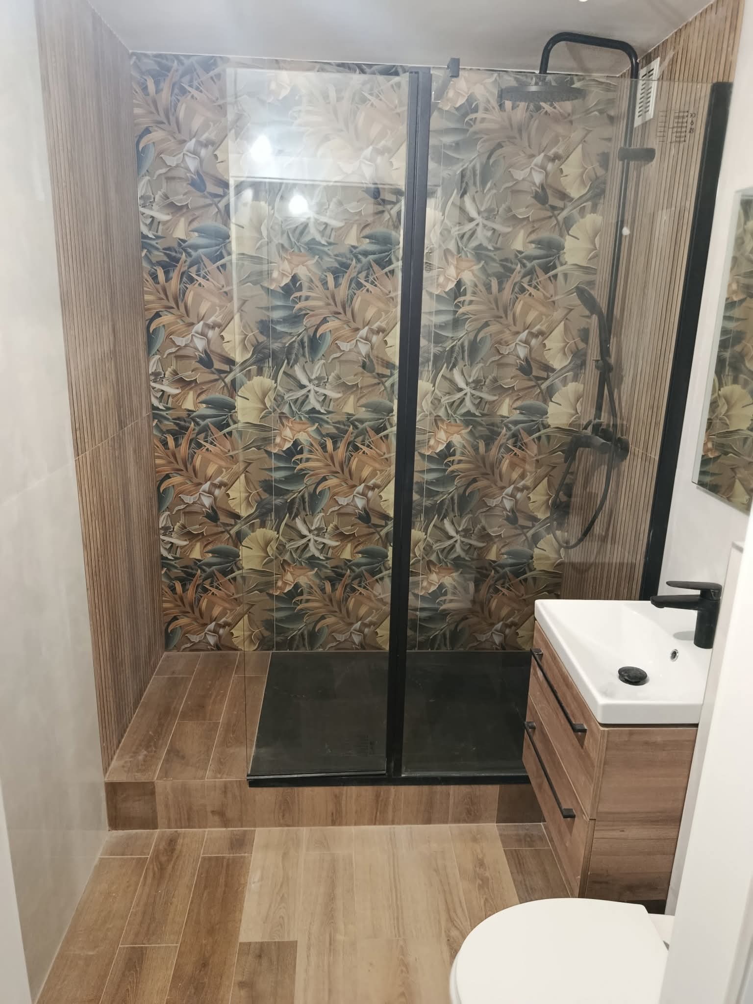 Nowoczesna łazienka po remoncie: kabina prysznicowa z wzorzystymi płytkami, drewnopodobna podłoga i szafka pod umywalką z czarną armaturą.