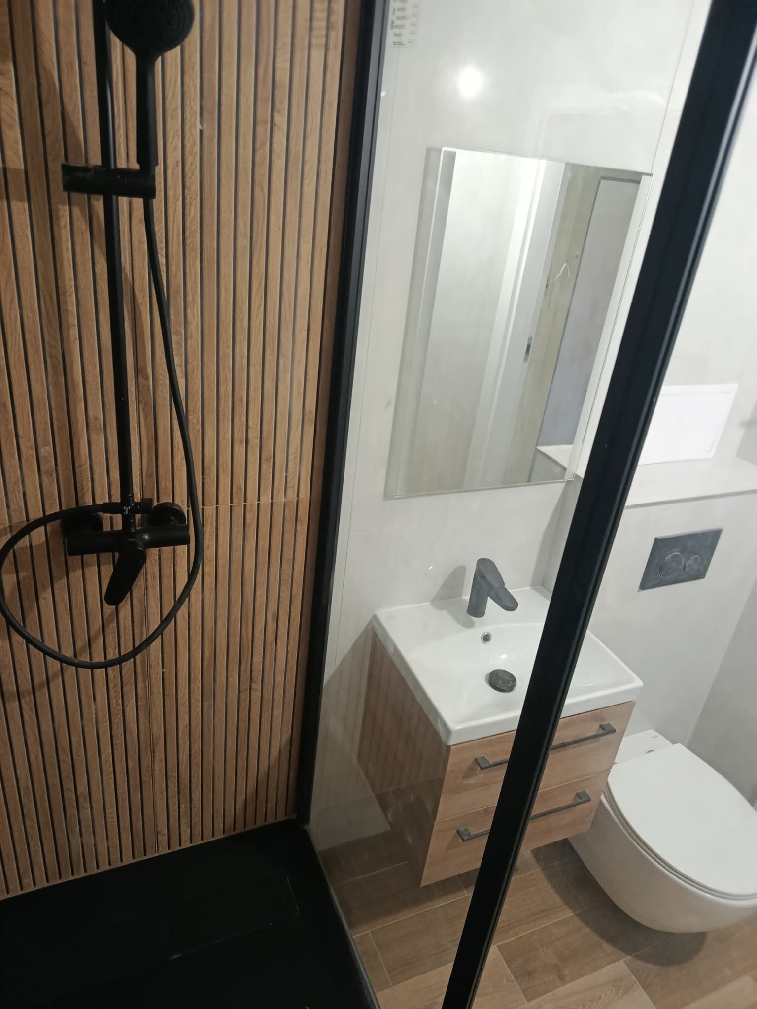 Nowoczesna łazienka z kabiną prysznicową typu walk-in, drewnianymi panelami ściennymi, czarną armaturą i szafką pod umywalką z szufladami. Minimalistyczny design.