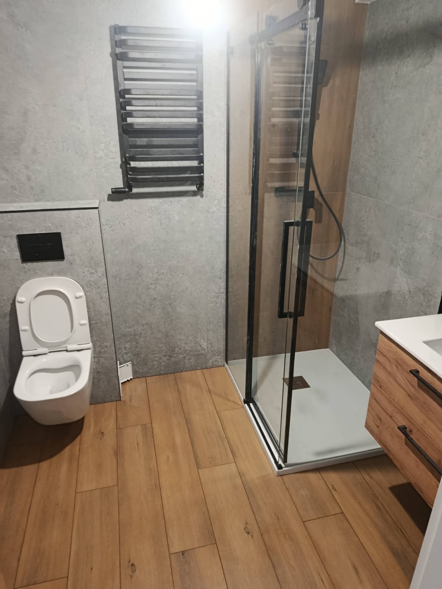 Nowoczesna łazienka z kabiną prysznicową typu walk-in, drewnopodobnymi płytkami na podłodze i szarymi płytkami na ścianach. Uwagę zwraca minimalistyczny grzejnik drabinkowy.